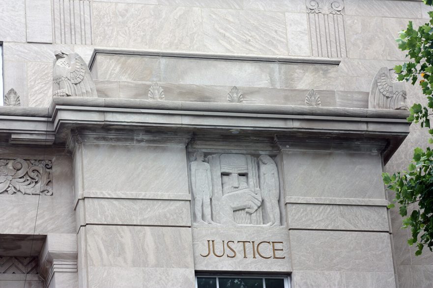 West façade, south corner, note the Art Deco carving of Justice, as well as the eagles and leaf that adorn the banding around the top of the building, circa 2022.